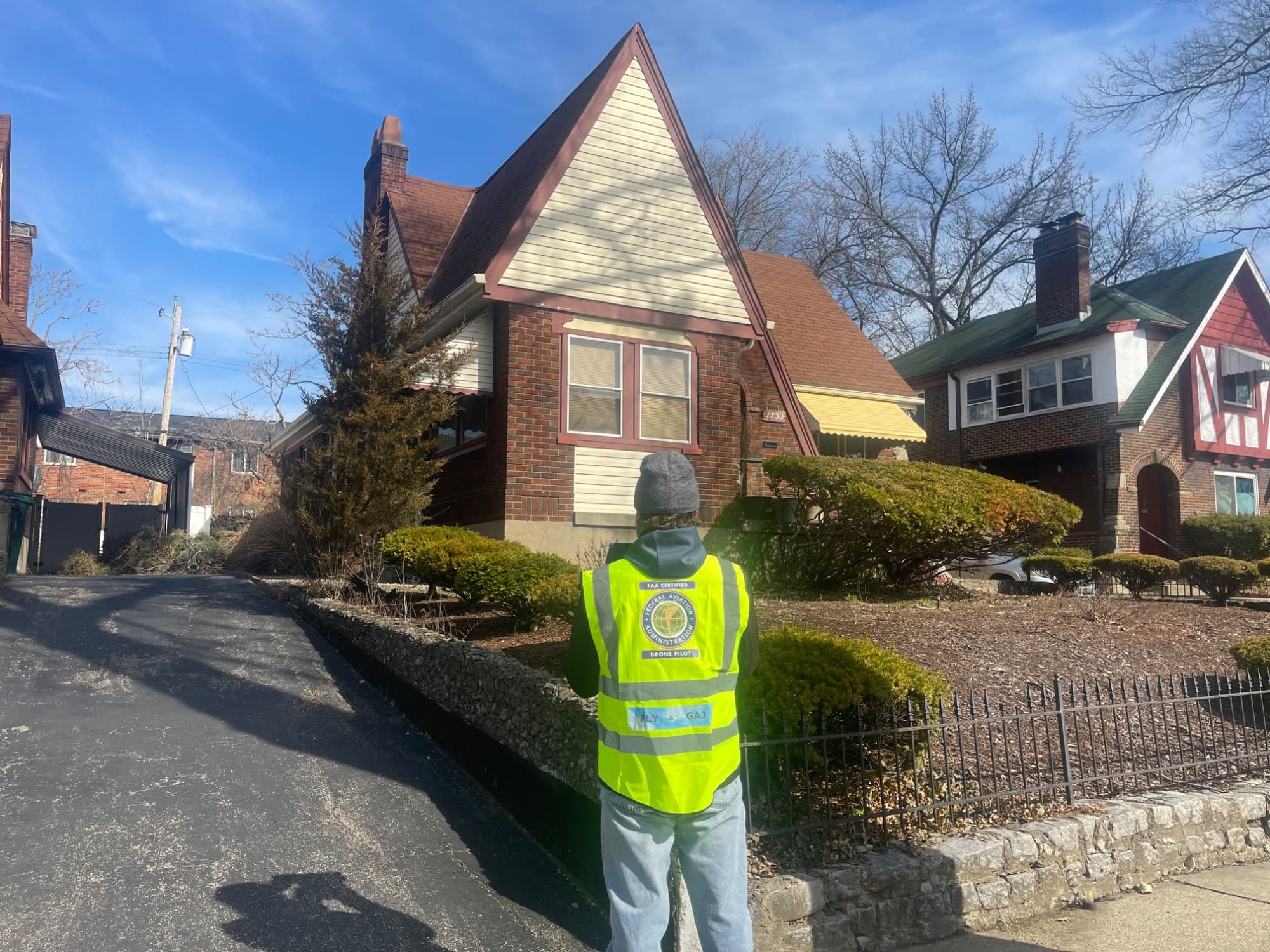 FAA-certified drone pilot in a high-visibility vest standing in front of a residential roofline during a field inspection
